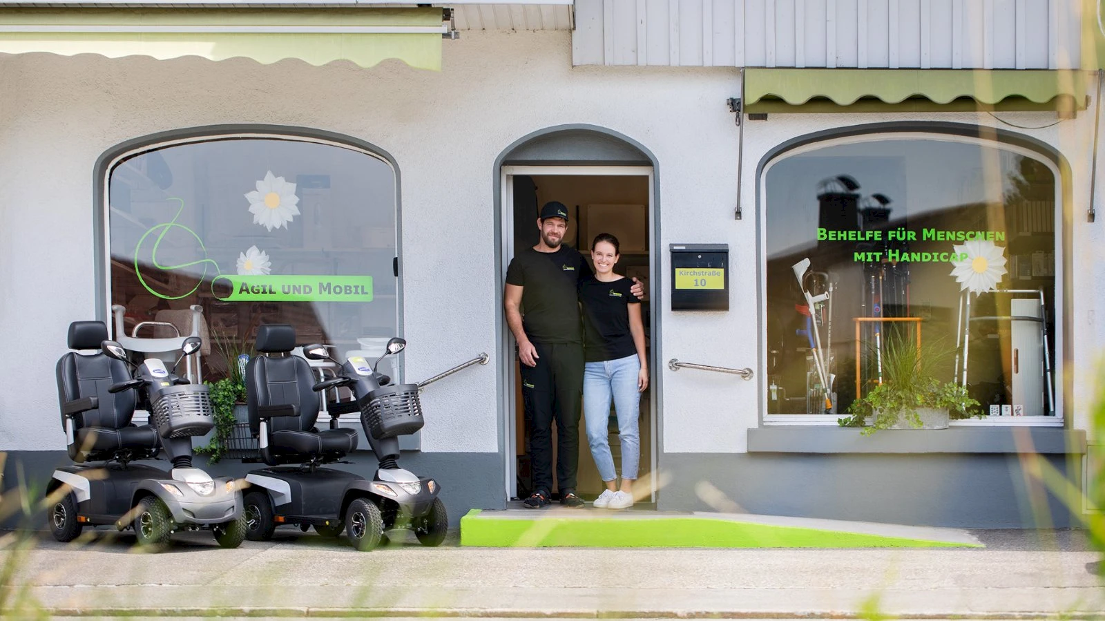 Franz-Josef und Melissa Feurstein stehen in der T&uuml;r ihres Gesch&auml;fts in der Kirchstra&szlig;e 10 in Lustenau, davor zwei Elektromobile